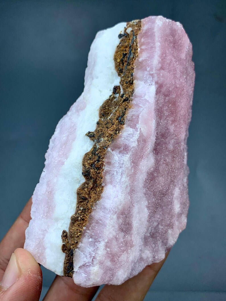 May include: A raw, natural mineral specimen featuring layers of pink, white, and brown hues. The pink sections have a textured appearance, while the white areas are smoother. A dark brown vein runs through the center of the stone.