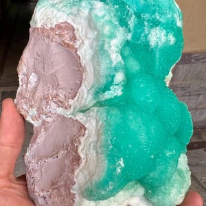 May include: A large, raw mineral specimen featuring a combination of colors and textures. The piece displays a vibrant turquoise-green, contrasted with areas of white and a muted pinkish-brown. The mineral's surface has a crystalline structure.