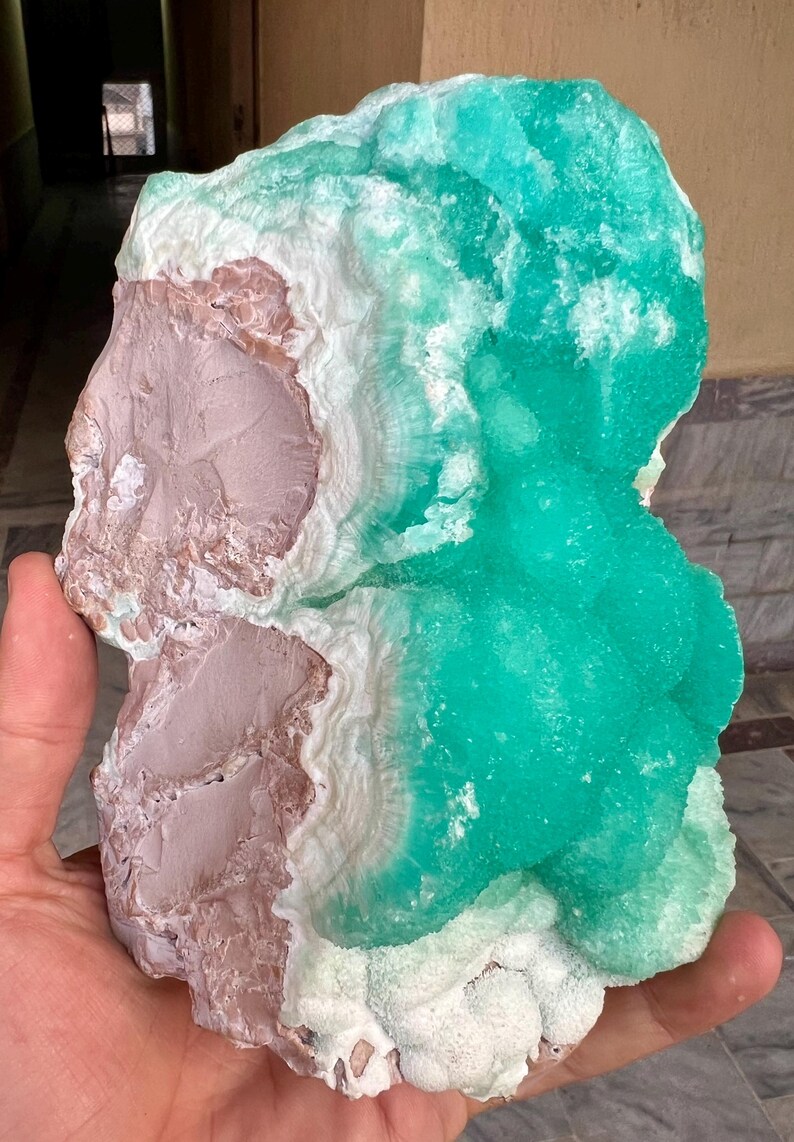 May include: A large, raw mineral specimen featuring a combination of colors and textures. The piece displays a vibrant turquoise-green, contrasted with areas of white and a muted pinkish-brown. The mineral's surface has a crystalline structure.