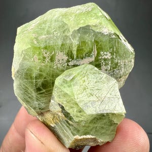 May include: A vibrant, translucent green crystal cluster. The mineral features a large, faceted main structure with a smaller, attached crystal. The surface has a textured appearance with subtle variations in color and some light-colored inclusions.
