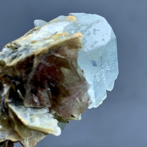 Peut inclure: Gros plan sur un cristal d'aigue-marine bleu p&acirc;le de forme hexagonale. Le cristal est partiellement fix&eacute; &agrave; une matrice de min&eacute;raux bruns et beiges, cr&eacute;ant un aspect brut. Le fond est un gris neutre flou.