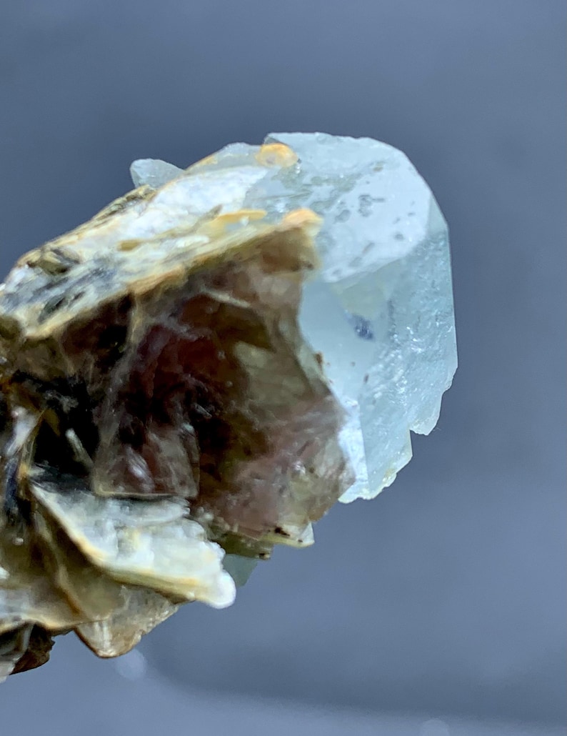 Peut inclure: Gros plan sur un cristal d'aigue-marine bleu p&acirc;le de forme hexagonale. Le cristal est partiellement fix&eacute; &agrave; une matrice de min&eacute;raux bruns et beiges, cr&eacute;ant un aspect brut. Le fond est un gris neutre flou.