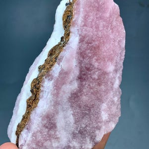 May include: A large, rough-hewn mineral specimen with a prominent pink hue. The stone features a white section and a brown, textured band. The overall appearance is natural and unpolished, showcasing the raw beauty of the mineral.