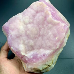 May include: A large, raw, pink mineral specimen with a bubbly, textured surface. The stone has a light pink hue with areas of white and tan. The overall shape is irregular and rough, showcasing natural formations.