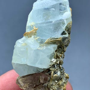 Peut inclure: Sp&eacute;cimen brut de cristal d'aigue-marine. Le cristal bleu p&acirc;le est partiellement transparent et repose sur une matrice de min&eacute;raux bruns et dor&eacute;s. La forme brute et naturelle de la pierre pr&eacute;cieuse est visible, mettant en valeur sa texture et sa structure uniques.