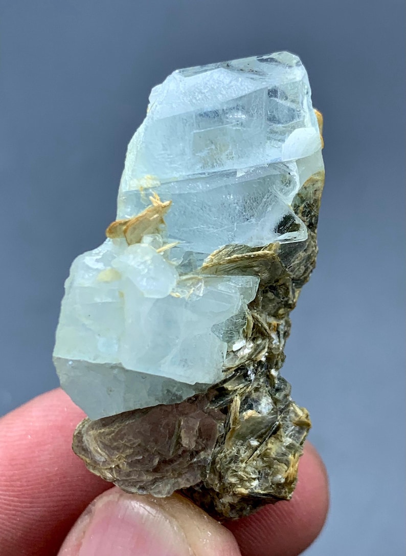 Peut inclure: Sp&eacute;cimen brut de cristal d'aigue-marine. Le cristal bleu p&acirc;le est partiellement transparent et repose sur une matrice de min&eacute;raux bruns et dor&eacute;s. La forme brute et naturelle de la pierre pr&eacute;cieuse est visible, mettant en valeur sa texture et sa structure uniques.