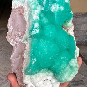 May include: A large, raw mineral specimen featuring a vibrant, teal-green crystalline formation atop a base of pale pink and white stone. The rough-hewn edges and natural textures highlight the unique geological composition of this specimen.