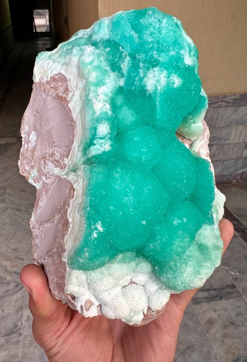 May include: A large, raw mineral specimen featuring a vibrant, teal-green crystalline formation atop a base of pale pink and white stone. The rough-hewn edges and natural textures highlight the unique geological composition of this specimen.