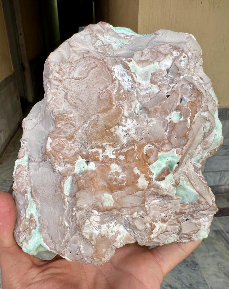 May include: A large, irregularly shaped rock with a mix of colors. The rock has a light brown base with areas of white, green, and darker brown. The surface is textured with various patterns and formations.
