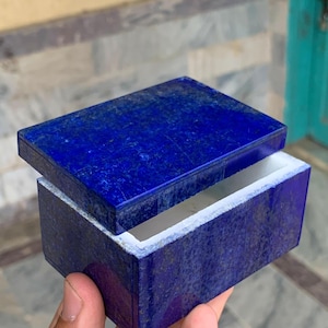 Puede incluir: Una caja rectangular hecha de piedra lapislázuli de color azul profundo con motas de pirita doradas visibles. La caja cuenta con una tapa a juego ligeramente desplazada, revelando un borde interior blanco en la parte superior de la base.