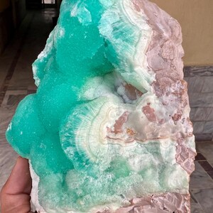 May include: A large, raw mineral specimen featuring a vibrant turquoise-green hue on one side, transitioning to white and pale pink on the other. The crystalline structure displays concentric patterns and a rough, natural texture. A unique decorative item.