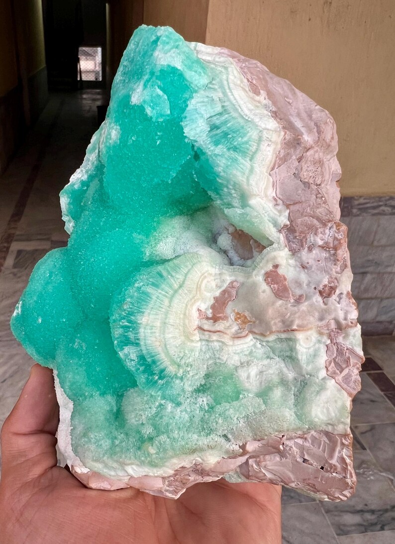 May include: A large, raw mineral specimen featuring a vibrant turquoise-green hue on one side, transitioning to white and pale pink on the other. The crystalline structure displays concentric patterns and a rough, natural texture. A unique decorative item.