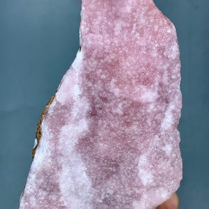 May include: A rough-cut, pink mineral specimen with a textured surface. The stone has areas of lighter pink and white, with a small section of brown and gold. The background is a solid, muted blue-gray.