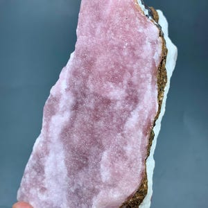 May include: A raw mineral specimen featuring a large, textured, pink crystalline formation. The pink mineral sits atop a white base with a brown and black edge. The specimen is held against a gray background.