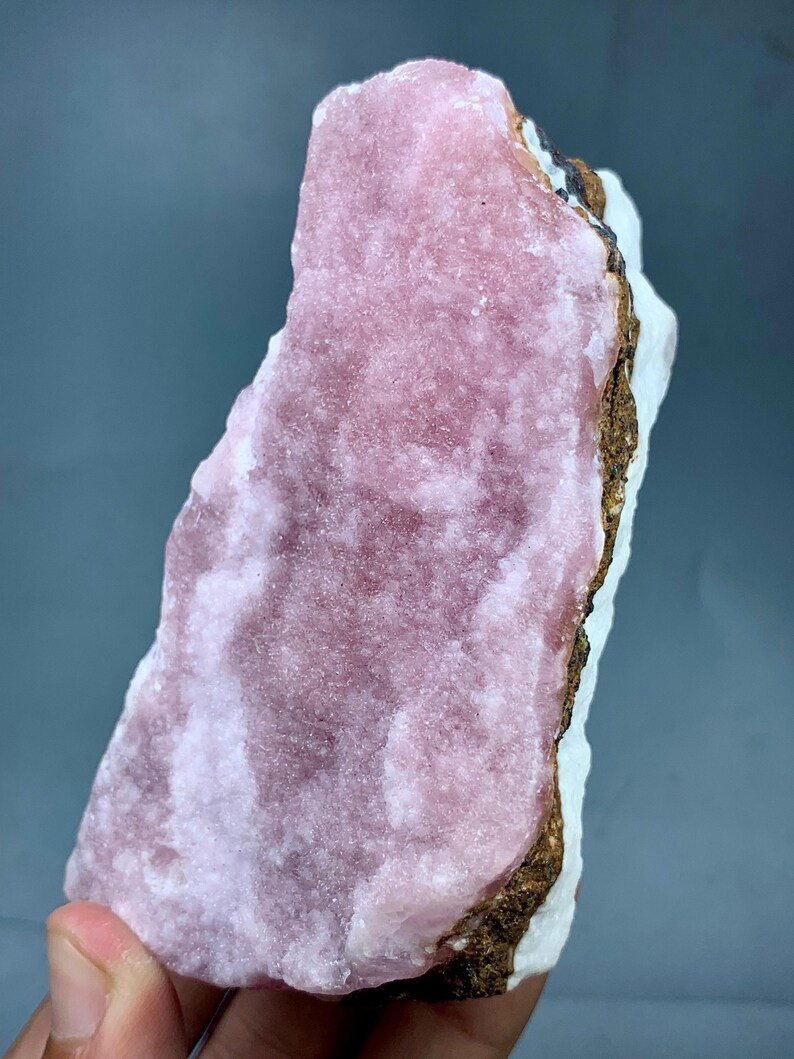 May include: A raw mineral specimen featuring a large, textured, pink crystalline formation. The pink mineral sits atop a white base with a brown and black edge. The specimen is held against a gray background.