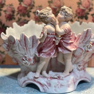 Puede incluir: Un cuenco de porcelana blanca y rosa con dos figuras esculpidas de pie frente a frente en el centro. El cuenco tiene un borde festoneado y texturizado con detalles florales en relieve. Las figuras visten prendas cortas y fluidas de tono rosa.
