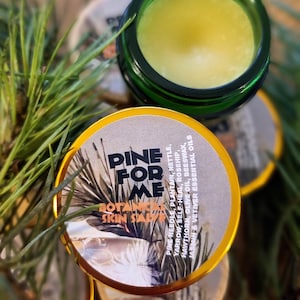 Op de afbeelding: Een groene pot gevuld met gele balsem, met de tekst "PINE FOR ME BOTANICAL SKIN SALVE" op het deksel. De ingrediënten zijn onder meer dennennaald, weegbree, brandnetel, duizendblad en essentiële oliën. Groene dennennaalden op de achtergrond.