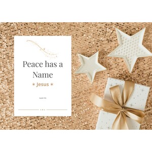 May include: A festive arrangement featuring a white gift box with a gold ribbon, two star-shaped decorative plates, and a card with the text "Peace has a Name * Jesus *" on a gold sequined background.