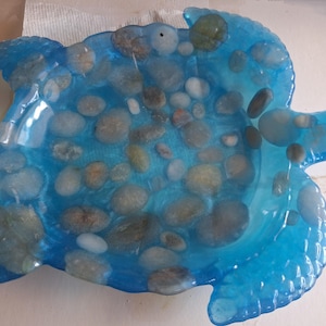 Peut inclure: Un plat artisanal en forme de tortue en résine bleue. Le plat est rempli de pierres lisses de différentes tailles, grises et blanches. La résine a une finition brillante, créant un effet aquatique. Le plat mesure environ 20 cm de long et 18 cm de large.