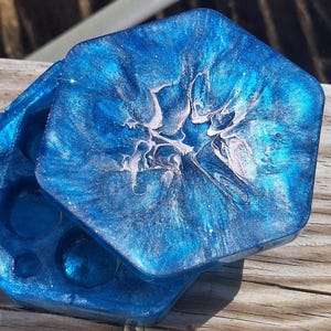 Könnte beinhalten: Sechseckiges blaues Untersetzer-Set aus Kunstharz mit schimmerndem Marmoreffekt. Die Untersetzer haben eine glänzende Oberfläche mit weißen und silbernen Akzenten. Das Set enthält einen Deckel und eine Basis mit kreisförmigen Vertiefungen.