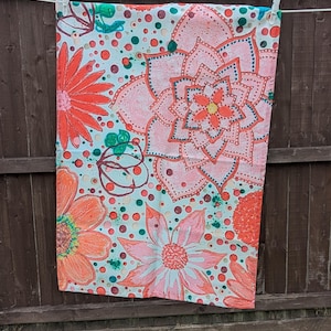 Könnte beinhalten: Ein Handtuch mit floralem Muster und hellblauem Hintergrund. Das Handtuch zeigt große rosa und orangefarbene Blumen mit grünen und roten Akzenten. Das Handtuch hängt an einer Wäscheleine vor einem Holzzaun.