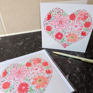 Könnte beinhalten: Zwei Grußkarten mit einem herzförmigen Blumendesign. Die Herzen sind mit roten, rosa und orangefarbenen Blumen und grünen Akzenten gefüllt. Ein Stift mit goldfarbener Spitze liegt auf der Oberfläche neben den Karten. Die Karten liegen auf einer dunklen, gesprenkelten Oberfläche.