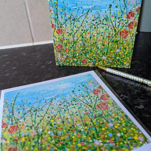 May include: Two art prints featuring a colorful floral meadow scene under a blue sky. The artwork includes red flowers, green stems, and a variety of colorful dots. A pen rests near the bottom print.