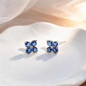 May include: A pair of silver stud earrings, each featuring a four-petal flower design. Each petal is composed of a deep blue gemstone. The earrings are displayed on a white, organically shaped ceramic dish, with a soft, neutral-toned fabric in the background.