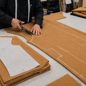 May include: A person in a black shirt uses metal scissors to cut a large piece of tan fabric. A white paper pattern labeled FRONT CUT - 2 rests on a stack of identical tan fabric pieces, while white chalk outlines are visible on the main fabric.
