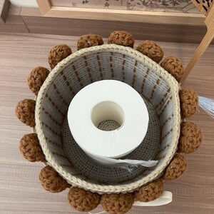 May include: A woven toilet paper holder with a cream-colored rim and a ring of brown crocheted balls around the top. The holder contains a roll of white toilet paper. The holder sits on a light brown surface.