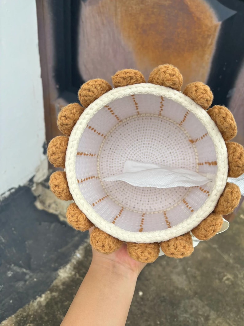 May include: A handmade, round tissue box cover. The cover is white with a crocheted border and brown pom-poms around the edge. The interior is white, and a tissue is visible inside. The cover is held by a hand.
