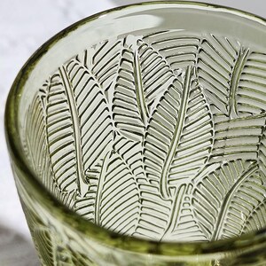 May include: Close-up of a green-tinted glass with a leaf pattern etched into the interior. The glass has a thick rim and a textured surface, showcasing the intricate design. The leaf pattern is repeated throughout the glass.