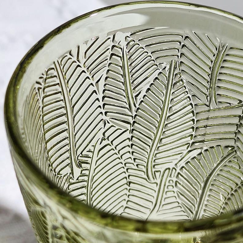 May include: Close-up of a green-tinted glass with a leaf pattern etched into the interior. The glass has a thick rim and a textured surface, showcasing the intricate design. The leaf pattern is repeated throughout the glass.