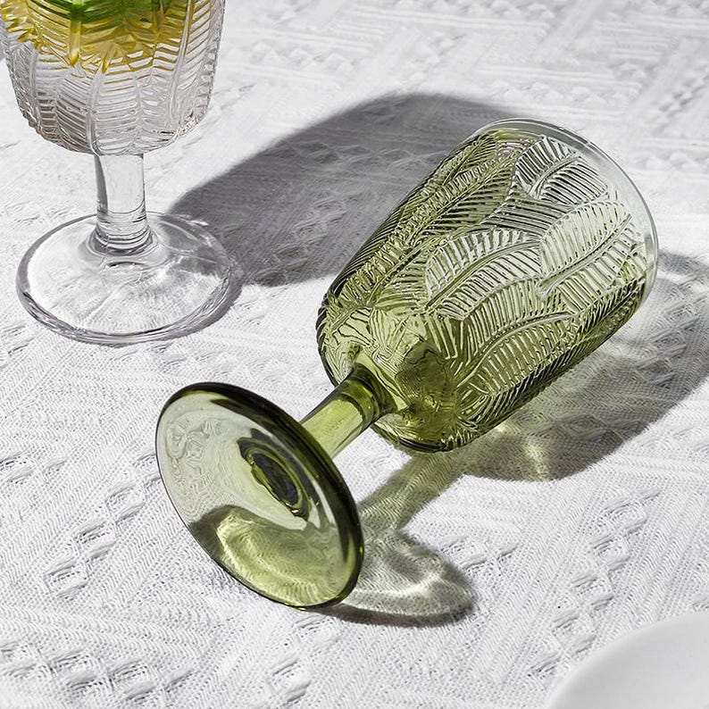 May include: Two stemmed glasses, one clear and one olive green, rest on a white textured surface. The green glass is lying on its side, showcasing a leaf-like pattern etched into the glass. The clear glass is upright, partially filled with a yellow liquid.