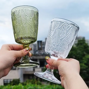 Puede incluir: Dos copas de vino con un patrón de hojas. Una copa es verde, la otra es transparente. Las copas se sostienen contra un fondo borroso de follaje verde y un edificio.