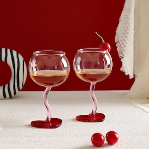 May include: Two clear wine glasses with a unique design. The stems are a light pink, curving upwards to support the round bowl. The base is a heart shape in red. One glass has a cherry on top. The glasses are filled with a reddish liquid.
