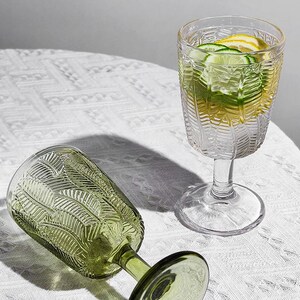 May include: Two stemmed glasses with a leaf pattern. One glass is olive green and lying on its side. The other is clear and filled with water, lemon, and cucumber slices. The glasses are on a white, textured surface.