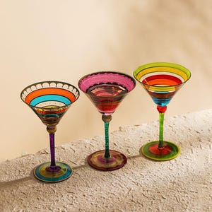 May include: Three colorful martini glasses with striped designs. Each glass features a different color scheme, including orange, blue, and green. The stems and bases are also decorated with vibrant colors, creating a unique and artistic look.