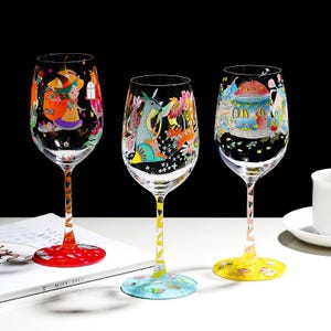 May include: Three colorful wine glasses with whimsical illustrations. Each glass features a different design, including a witch, a mythical creature, and a fairy house. The stems have patterned designs, and the bases are red, yellow, and blue.