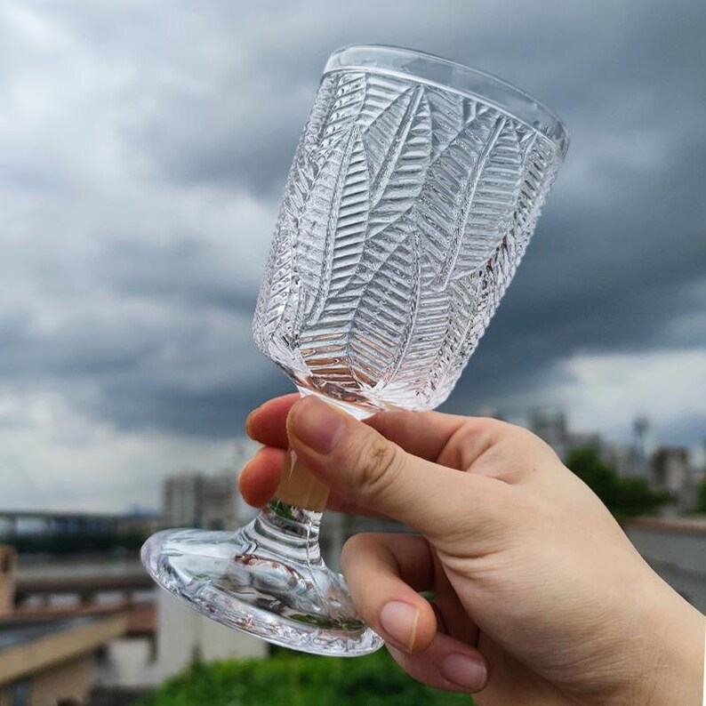 May include: A clear, stemmed glass with a textured, leaf-like pattern. The glass has a wide bowl and a slender stem. The background is a cloudy sky and a cityscape.
