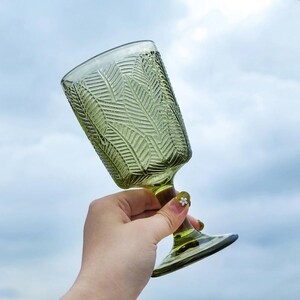 May include: A green-tinted glass goblet with a textured leaf pattern. The stem and base are also green. The glass is held against a cloudy sky background. The goblet is likely used for serving beverages.