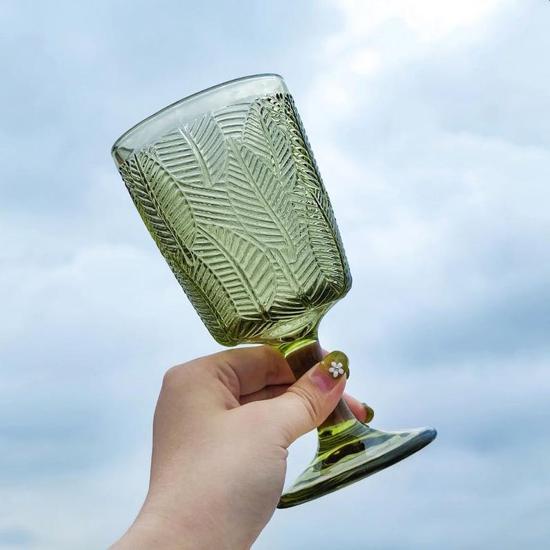 May include: A green-tinted glass goblet with a textured leaf pattern. The stem and base are also green. The glass is held against a cloudy sky background. The goblet is likely used for serving beverages.