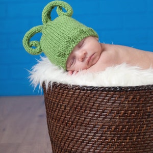May include: A newborn baby wearing a green knitted hat with two ear-like loops, sleeping in a brown wicker basket lined with white fur. The background is a bright blue wall.