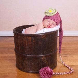 May include: A newborn baby sleeping in a wooden barrel wearing a pink and green knitted hat with a flower on top. The hat has a long tail that is tied with a pink pom-pom.
