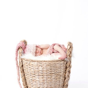 May include: A newborn baby wearing a pink and white knitted hat and shorts, sleeping peacefully in a woven basket lined with white faux fur. The basket has a braided handle and is set against a white background. The text "Twice As Nice Photography" is in the top left corner.