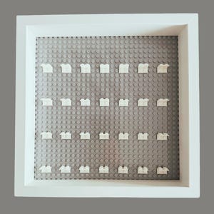May include: A white framed display case with a gray base, designed to hold small building blocks. The case features a grid of small circular studs, with several white building blocks attached. The frame is square and has a clean, modern aesthetic.
