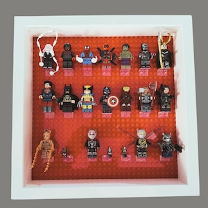 May include: A framed display of various superhero Lego minifigures. The figures are arranged on a red Lego baseplate within a white frame. Characters include Spider-Man, Batman, and Wolverine.