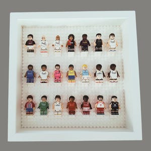 Może przedstawiać: Oprawiona ekspozycja z 18 miniaturowymi figurkami piłkarzy. Każda figurka ubrana jest w inny strój drużyny, o różnych kolorach i wzorach. Figurki ułożone są na białej podstawie w białej ramie.