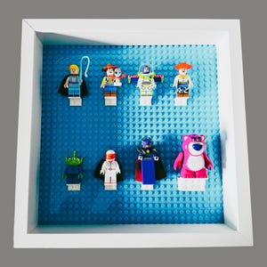 Può includere: Una vetrina con cornice bianca con varie minifigure Lego di Toy Story su una base Lego blu. La collezione comprende personaggi come Woody, Buzz Lightyear e Lotso. La cornice è quadrata e lo sfondo è grigio.