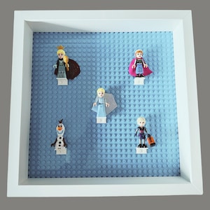 May include: A white framed display featuring five Lego figures from the movie Frozen. The figures are arranged on a light blue Lego baseplate. The figures include Elsa, Anna, Olaf, and Kristoff.